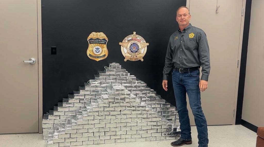 Sheriff Jason Ard standing beside a pyramid of tinfoil-wrapped methamphetamine bricks
